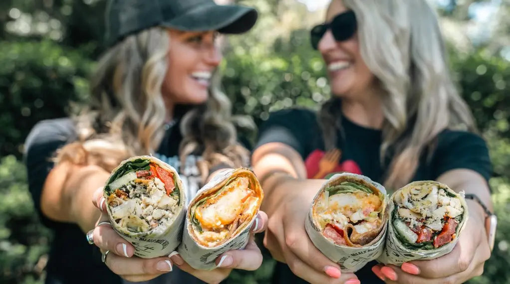 Two smiling women holding up stuffed wraps outdoors — a delicious and satisfying diet-friendly meal made to share.