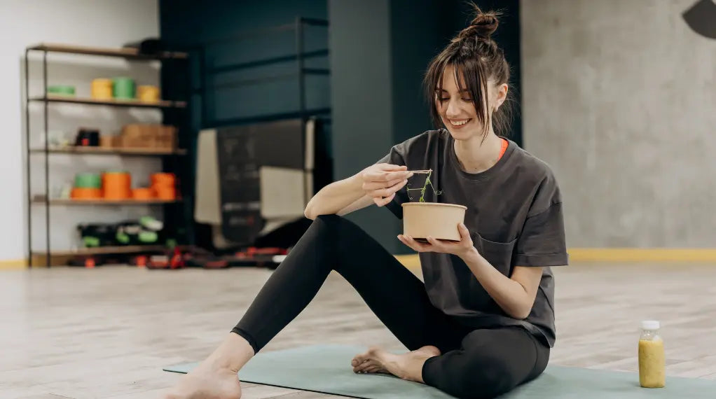 Woman eating clean, enjoying a container of prepared food while exercising on a yoga mat at home
