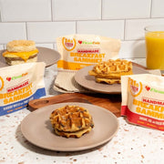 Handmade breakfast sandwiches with waffles and a glass of orange juice on a countertop.