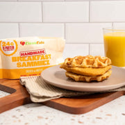 Waffles on a plate with a glass of orange juice and a package labeled 'Handmade Breakfast Sammies' on a white countertop.