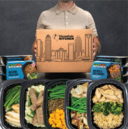 Clean Eatz Kitchen Basics Box — person holding branded shipping box with skyline graphic, surrounded by retail sleeves (Basic Ropa Vieja & Rice; Basic Beef & Sweet Potatoes) and four basic meal trays featuring chicken, steak strips, green beans, mashed potatoes, roasted potatoes, broccoli, brown rice, and diced sweet potatoes.