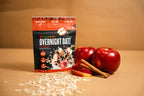 A bowl of overnight oats topped with apple slices beside a Clean Eatz Overnight Oatz Apple Cinnamon pouch, with whole apples and oats scattered on a tan surface.