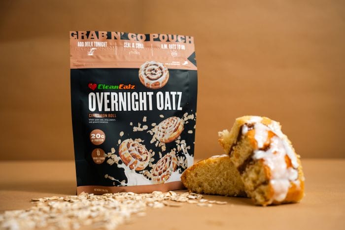 Clean Eatz Overnight Oatz Cinnamon Roll pouch with an iced cinnamon-roll bite in front and oats scattered on a tan background.