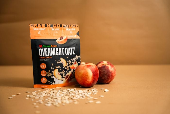 Clean Eatz Overnight Oatz Peaches & Cream pouch standing with fresh peaches nearby and oats sprinkled on a tan background.
