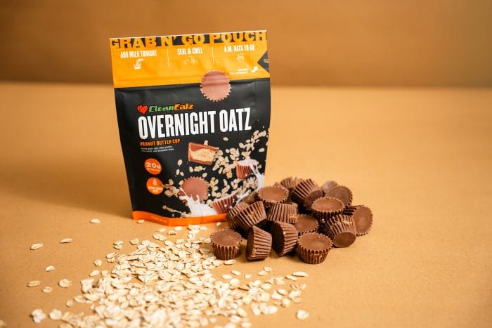 Clean Eatz Overnight Oatz ‘Peanut Butter Cup’ grab-n’-go pouch on a tan background with rolled oats and mini peanut butter cups scattered in front.