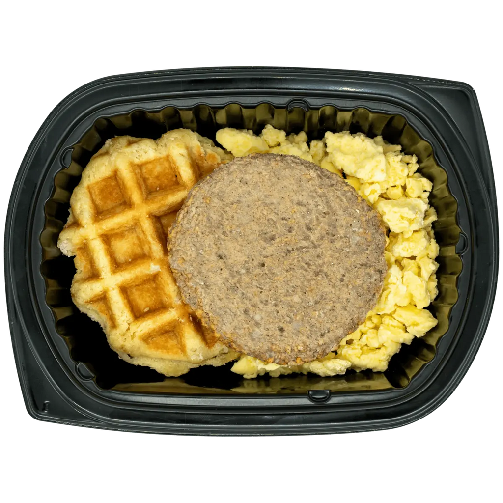 A breakfast meal in a black plastic clamshell container, featuring a golden-brown waffle, a round brown sausage patty, and scrambled yellow eggs on a white background.