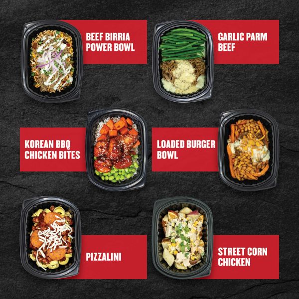 Six Clean Eatz Kitchen entrées on a black stone background — Beef Birria Power Bowl, Garlic Parm Beef, Korean BBQ Chicken Bites, Loaded Burger Bowl, Pizzalini, and Street Corn Chicken — each shown in individual meal trays with red labels.