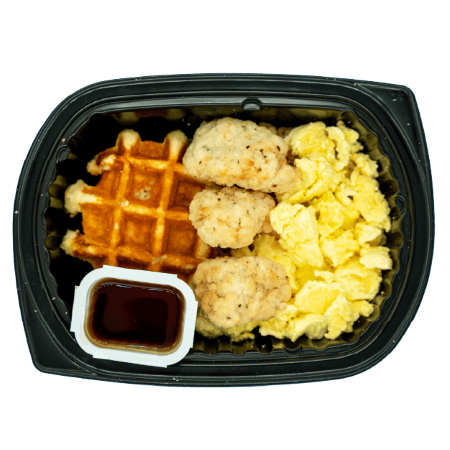 Chicken bites with a waffle, scrambled eggs, and a side cup of syrup in a black meal tray.