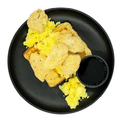 Chicken bites with scrambled eggs, a waffle, and a side cup of syrup on a black plate.