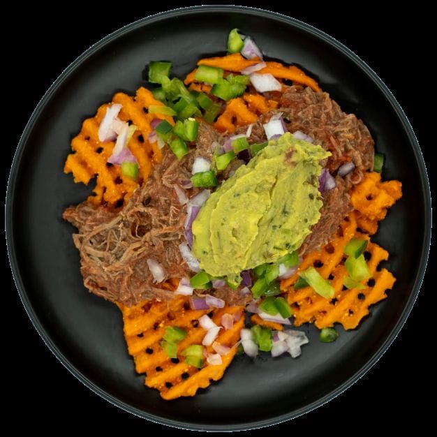 Shredded beef over sweet potato waffle fries topped with guacamole, red onions, and peppers on a black plate.