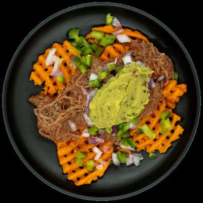 Shredded beef over sweet potato waffle fries topped with guacamole, red onions, and peppers on a black plate.