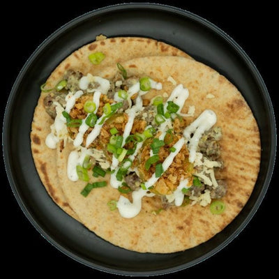 Pita topped with a creamy chicken mixture, shredded cheese, crispy onions, green onions, and white sauce on a black plate.