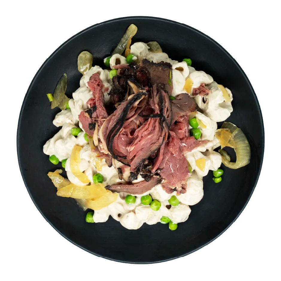 Prime rib over creamy pasta with mushrooms, onions, and green peas on a black plate.