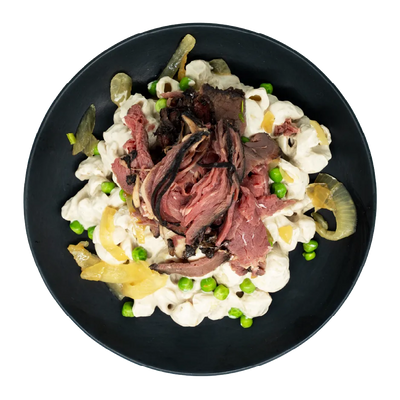 Prime rib over creamy pasta with mushrooms, onions, and green peas on a black plate.