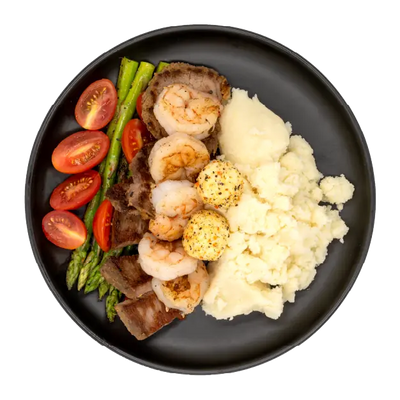Steak and shrimp with asparagus, tomatoes, mashed potatoes, and seasoned butter on a black plate.