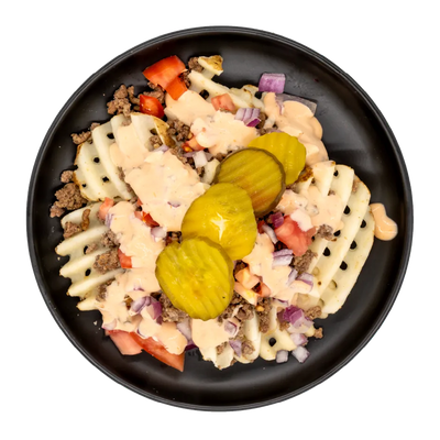 A top-down view of a loaded waffle fries dish in a black bowl, featuring creamy sauce, ground beef, diced tomatoes, red onion, and three pickle slices on a white background.