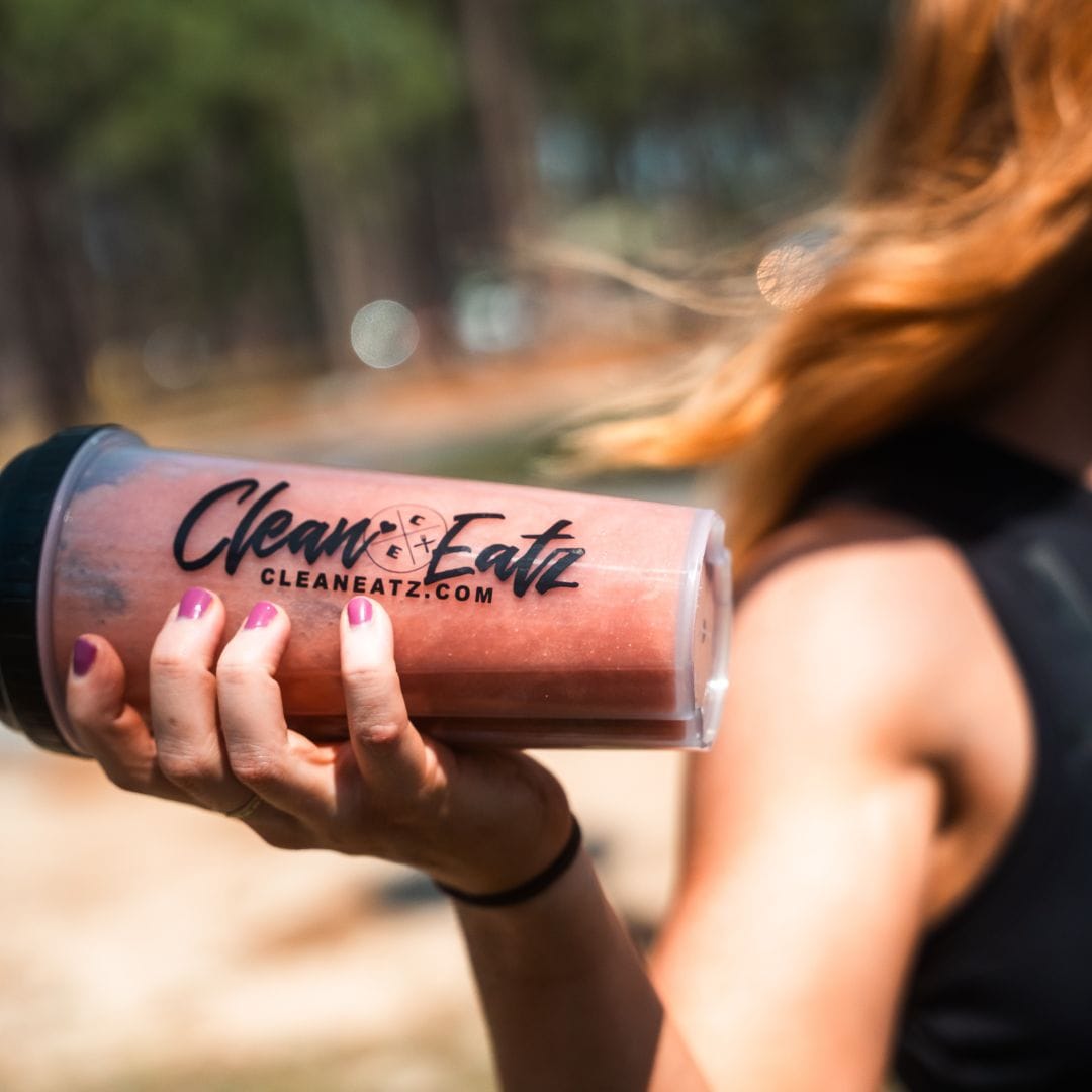 Person holding a pink smoothie cup with 'Clean Eatz' branding outdoors.