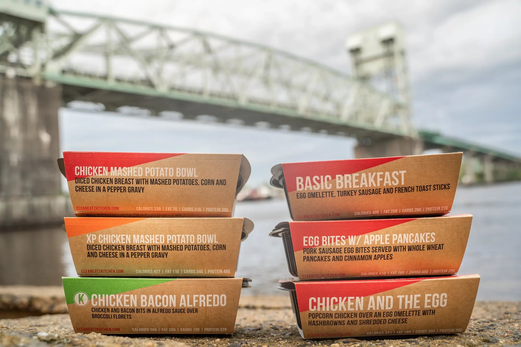 Stacks of food boxes with menu items on a waterfront background with a bridge.